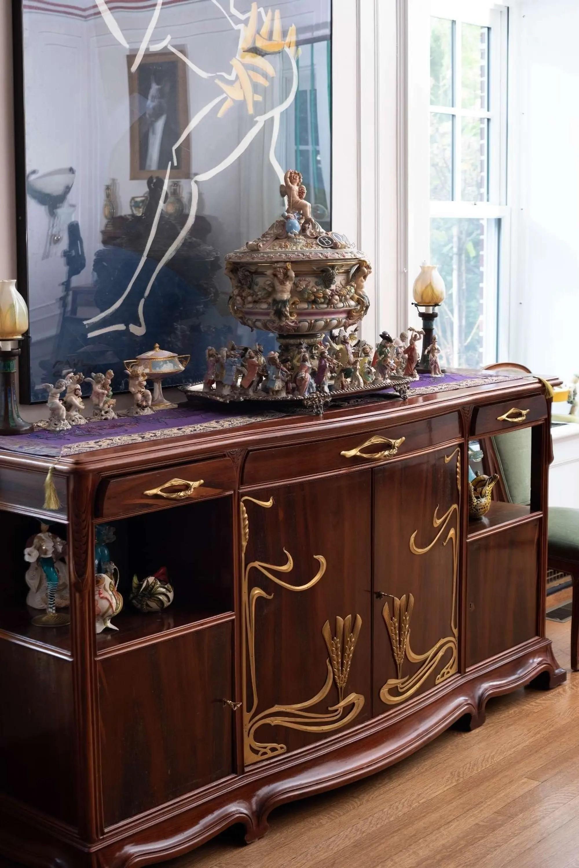 A dark wood Art Nouveau buffet with gold details, topped with an ornate lidded bowl, numerous figurines, and lamps, set beneath a large abstract artwork.