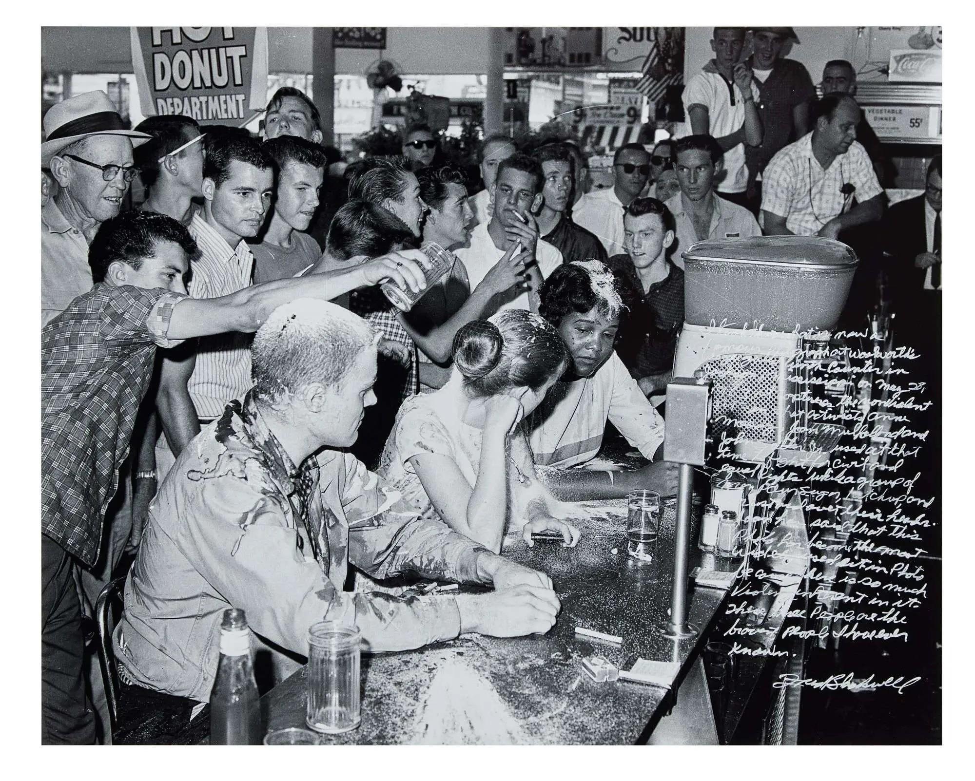 Black and white photo of a Civil Rights sit-in: Black and white protesters, covered in food, sit at a diner counter as hostile white men surround and assault them.