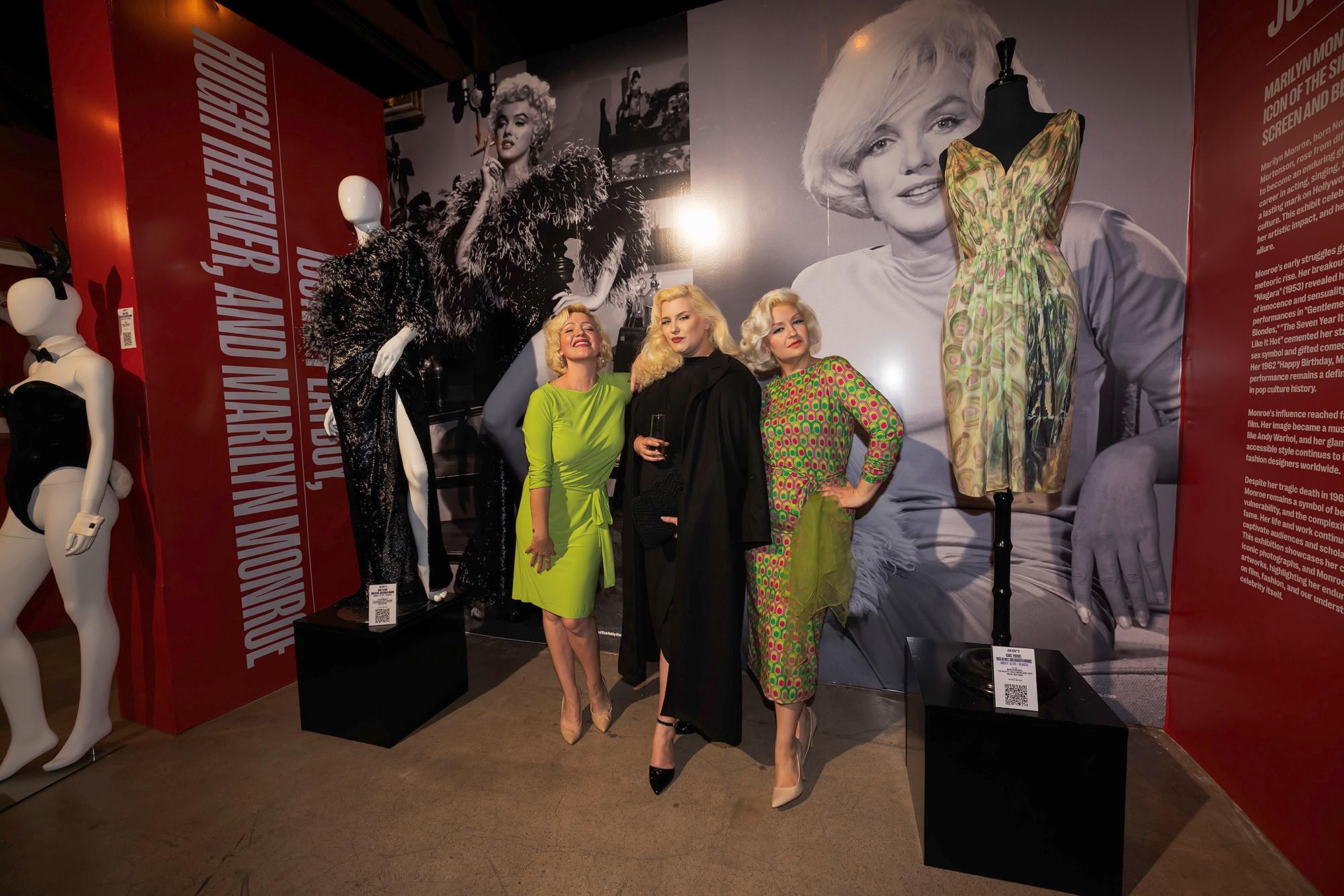 three blonde women are standing next to each other in front of a marilyn monroe poster .