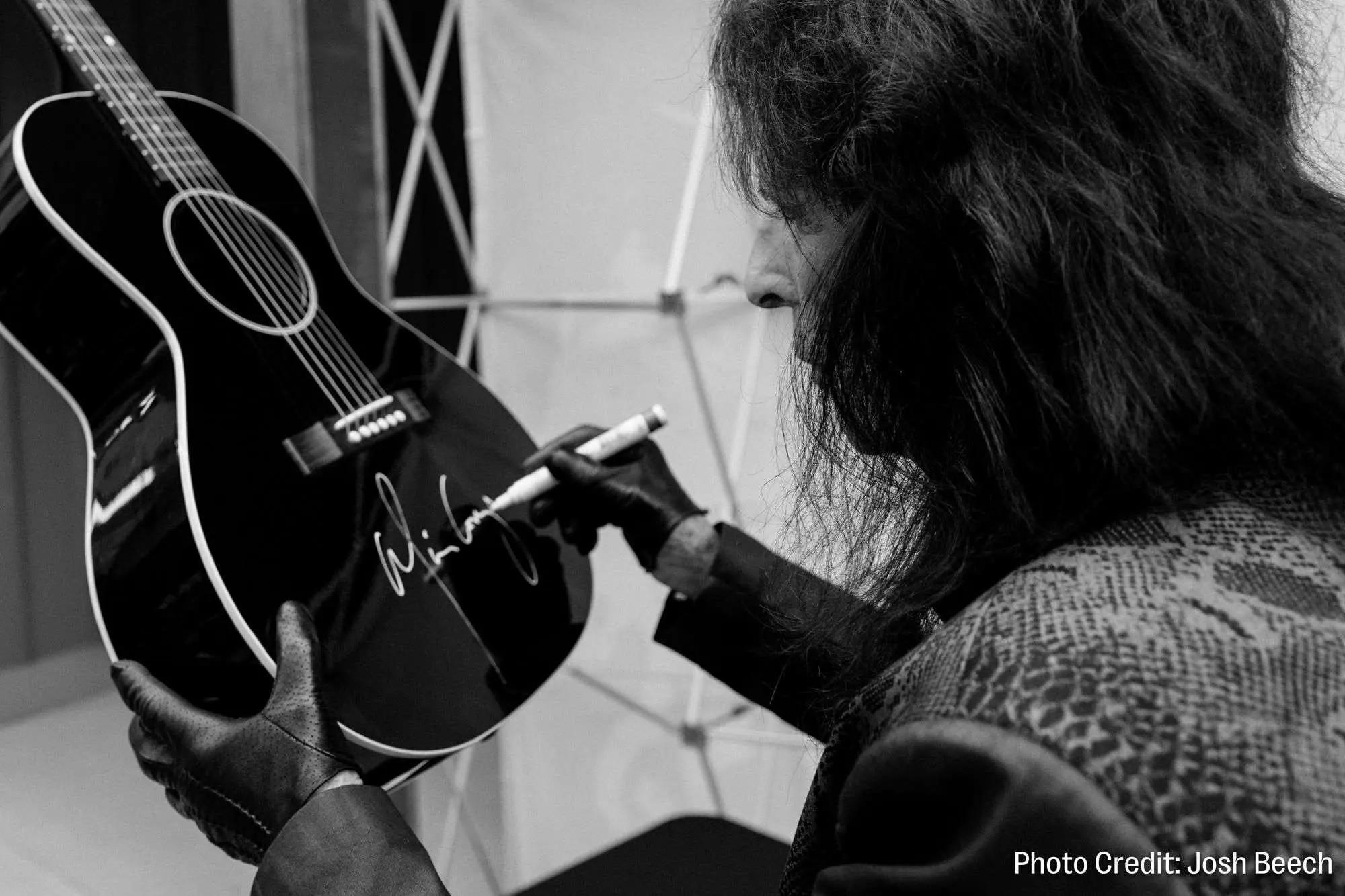 Black and white image of a person with long dark hair signing a black acoustic guitar.