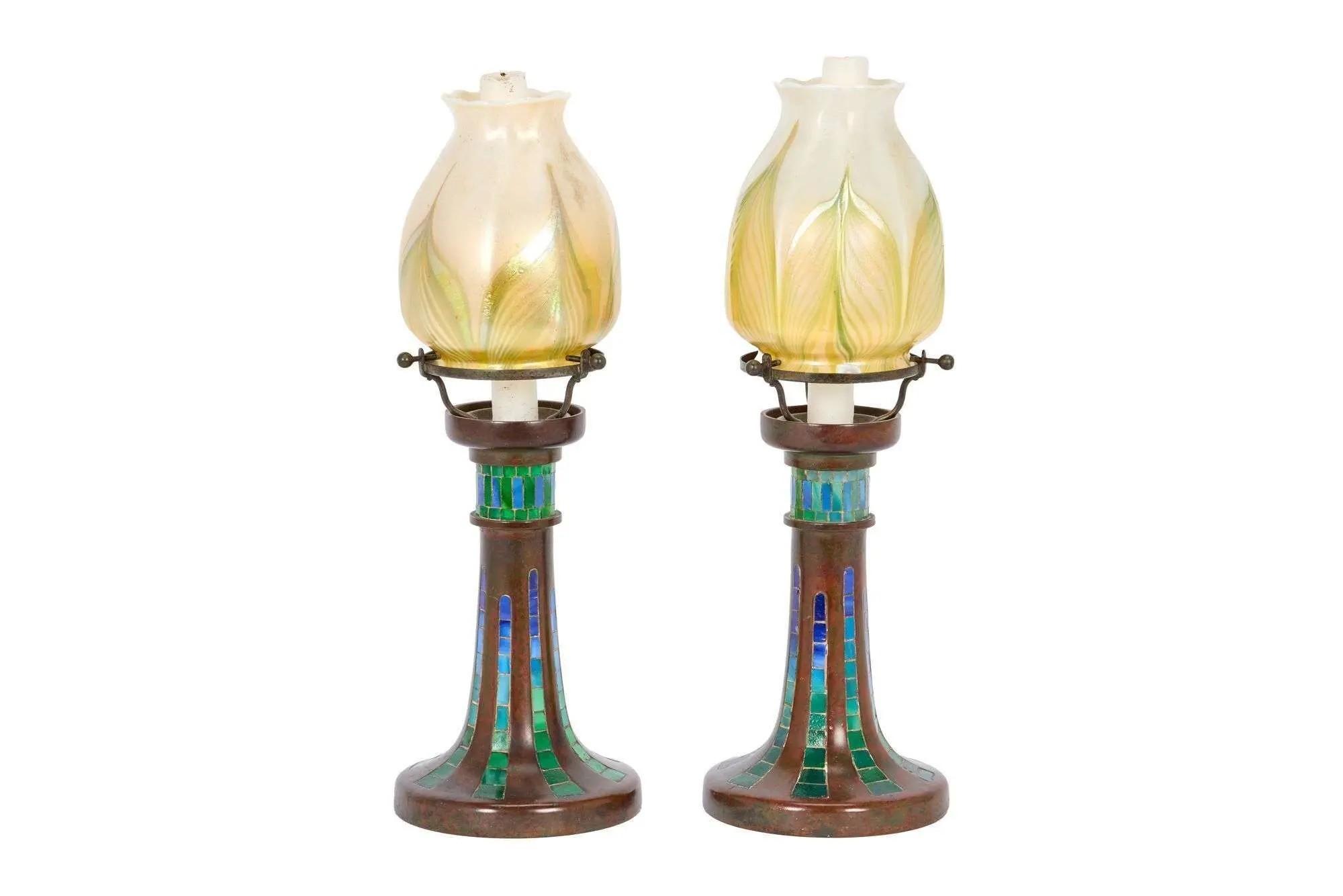 Lot #297. Whoopi Goldberg | Tiffany Studios Pair of Lighthouse Candlesticks A pair of table lamps with iridescent, leaf-patterned glass shades and bronze bases inlaid with blue and green mosaic tiles.