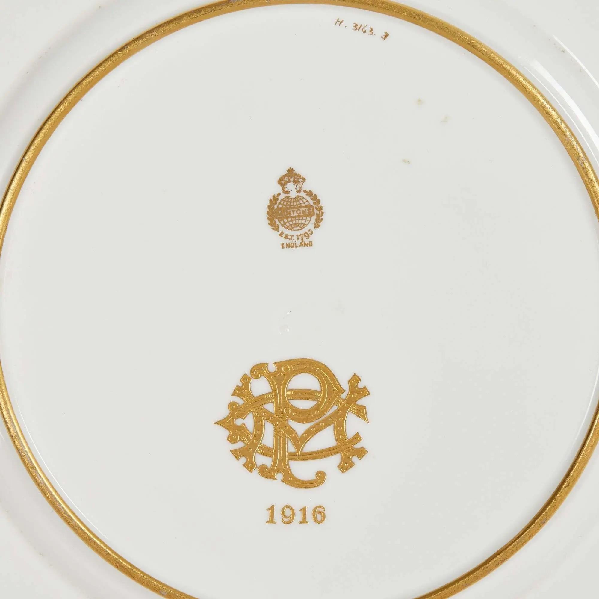 Back of a white ceramic plate with a gold rim, featuring a gold manufacturer's crest (EST. 1770 ENGLAND), a decorative gold monogram, and the year 1916.