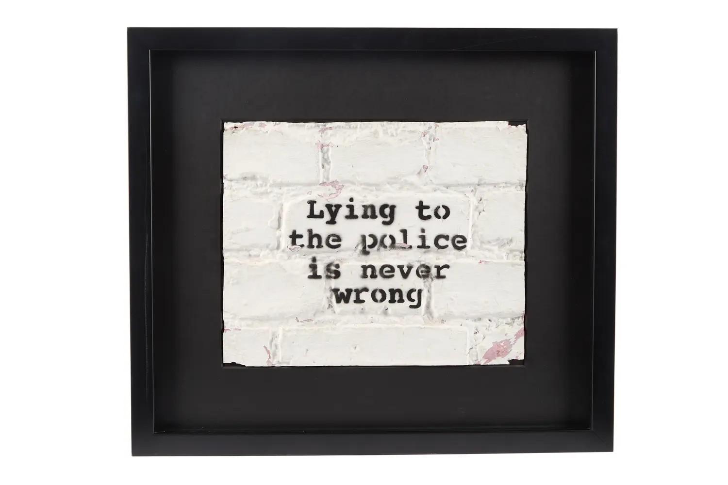 Lot #1. Banksy | "Lying to the Police is Never Wrong" Original 2001 Cargo Exhibition Painting (with Book) A framed artwork featuring the text "Lying to the police is never wrong" stenciled on a white brick wall texture.