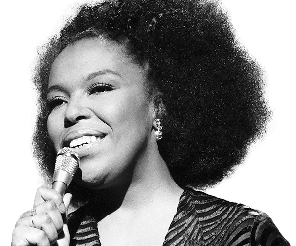 Two overlapping photos of a singer with curly hair and sunglasses at a keyboard, one in color and one in black and white, with a "Coming Soon" sticker.