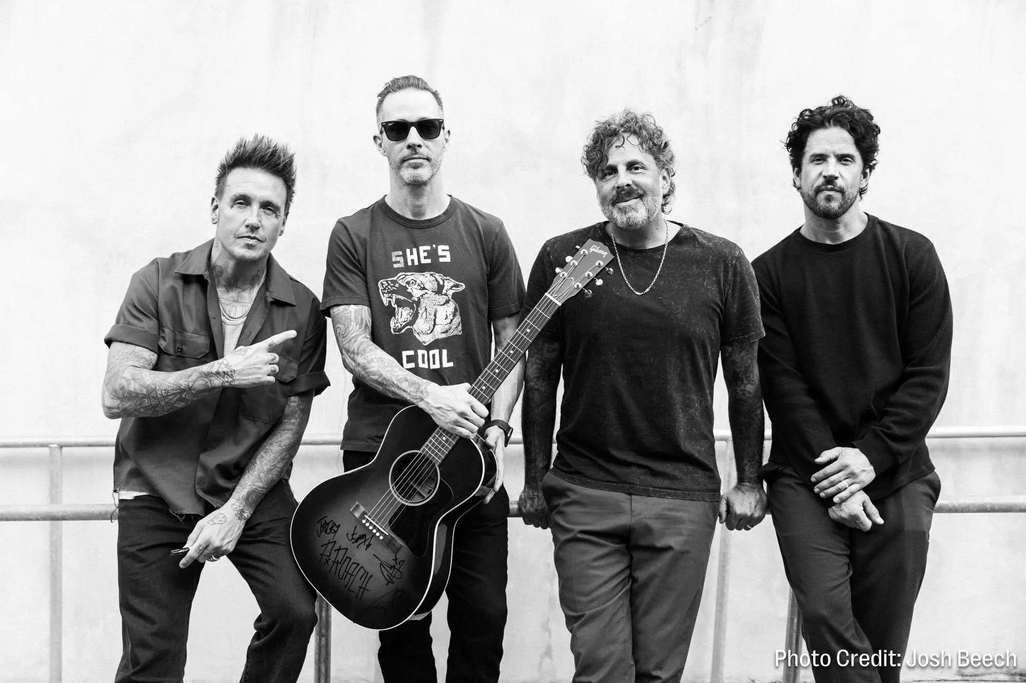 Black and white photo of four men, likely a band, posing together with one holding an acoustic guitar.