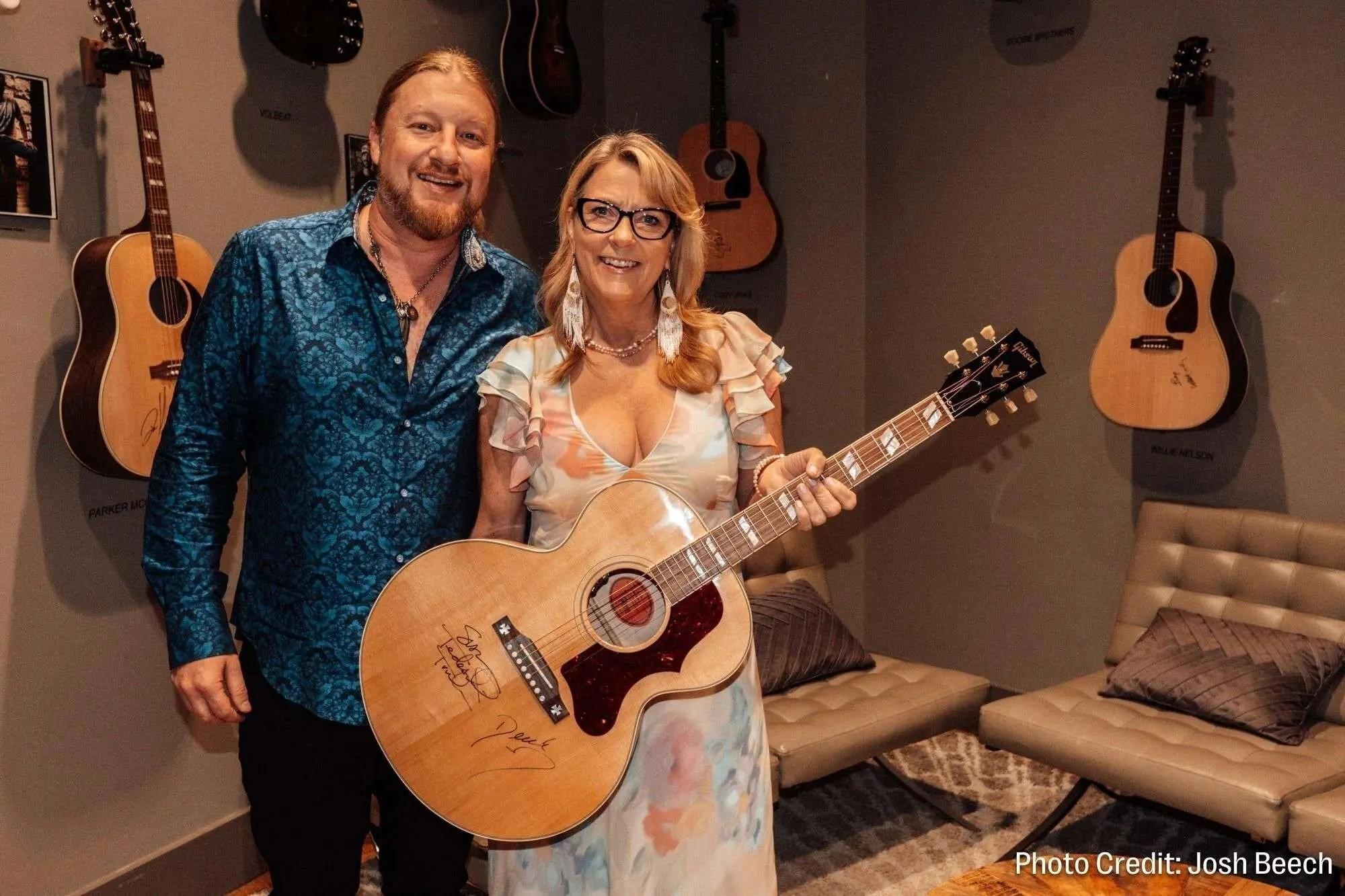 A smiling man and woman, with the woman holding a signed acoustic guitar, in a room lined with hanging guitars.