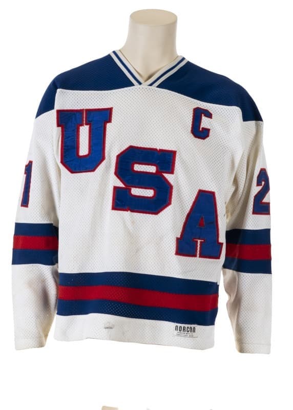 White "USA" hockey jersey with blue letters, a captain's "C", and red and blue stripes, displayed on a mannequin.