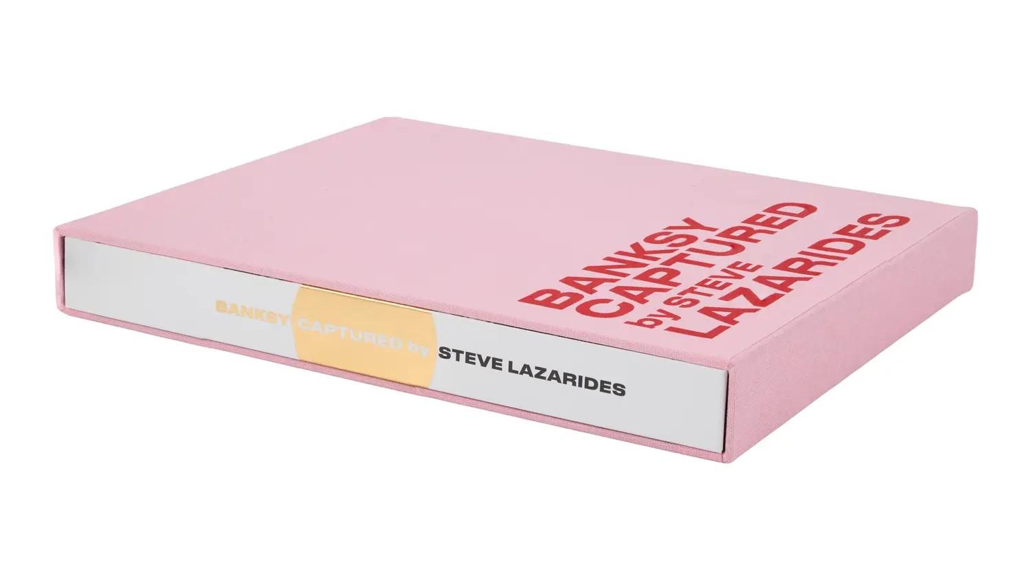 Lot #6. Banksy | Steve Lazarides Signed Limited-Edition "Banksy Captured - Vol. 2" Photo Book A pink book titled "Banksy Captured by Steve Lazarides" with red text on the cover and gold text on the spine.