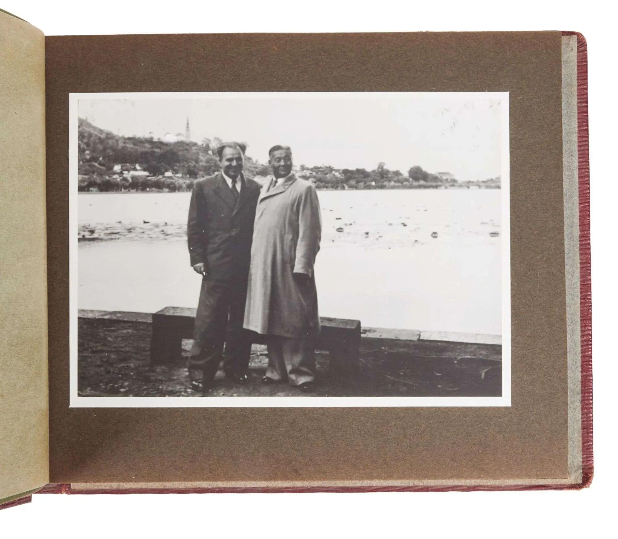 Black and white photo of two men standing by a river with a town and church steeple in the background, mounted in an album.