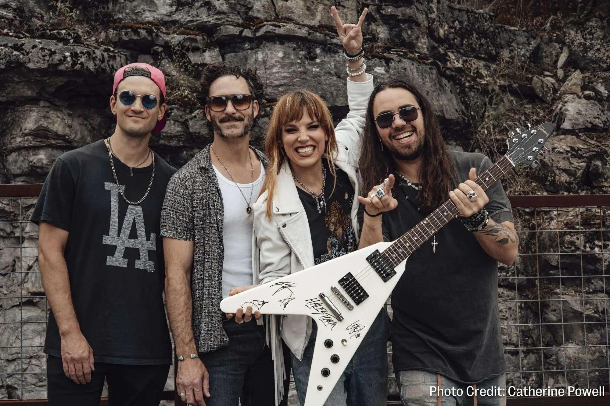 Four smiling rock band members pose with a white V-shaped guitar, one making a rock-on gesture.