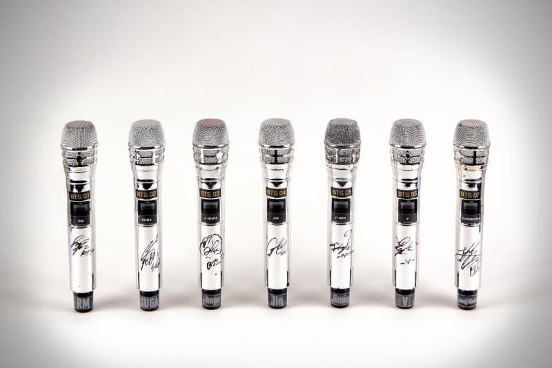 Seven silver microphones, each with a BTS member's signature and name, lined up on a white background.