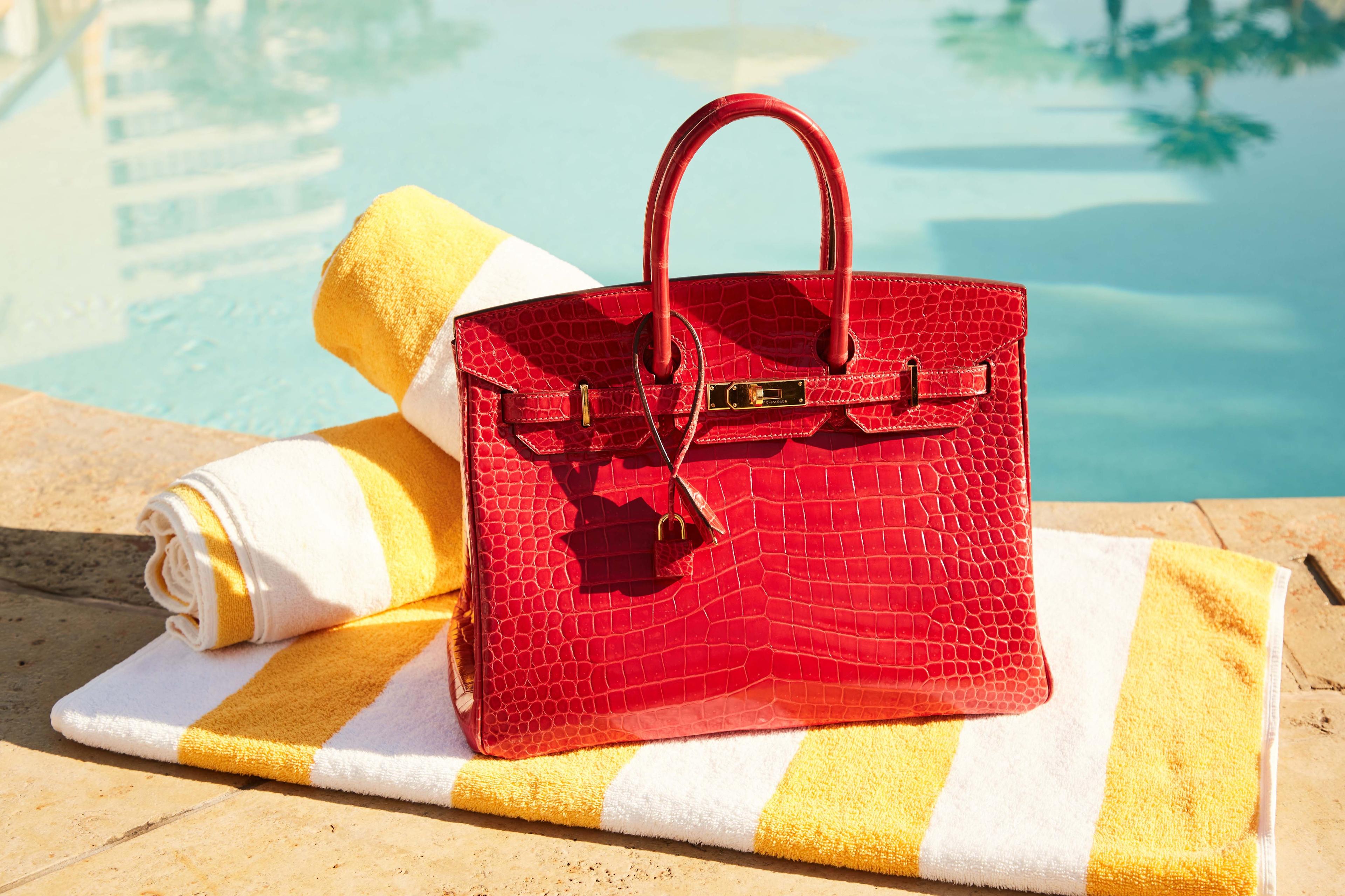 Lot 7, Hermes Red Birkin a red purse is sitting on top of a yellow and white striped towel next to a pool .