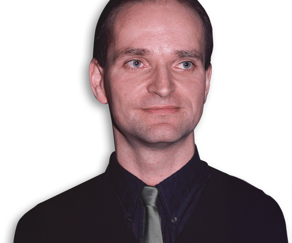 A man with short dark hair and different colored eyes wears a dark shirt, dark vest, and light grey tie, looking slightly to his left against a black background.