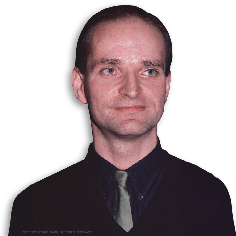 A man with short dark hair and different colored eyes wears a dark shirt, dark vest, and light grey tie, looking slightly to his left against a black background.