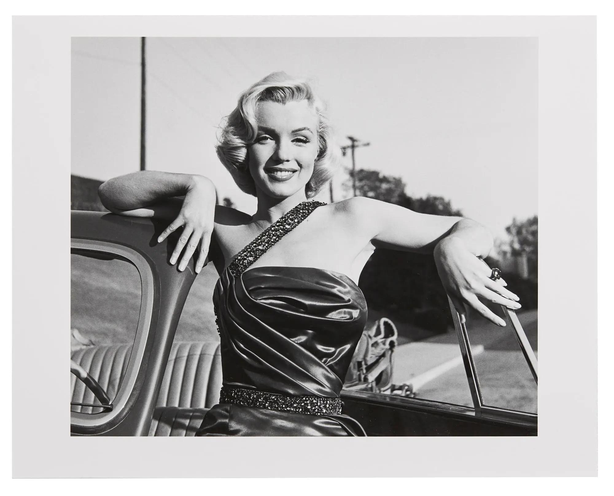 Lot #70. Marilyn Monroe | 1953/1990s Black and White Photograph by Frank Worth Black and white photo of Marilyn Monroe smiling, leaning on an open car window.