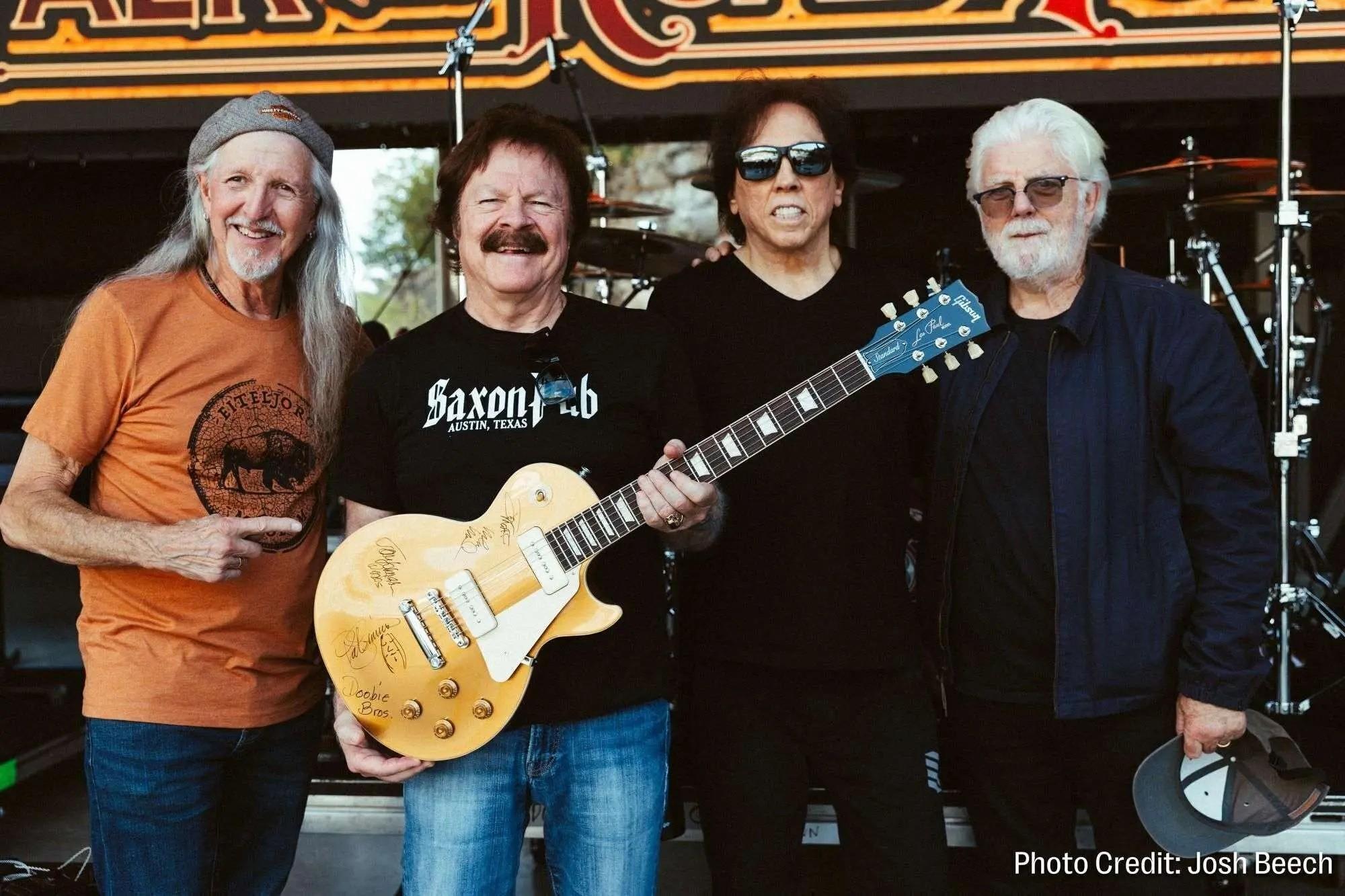 Four musicians pose together, one holding a signed yellow electric guitar.