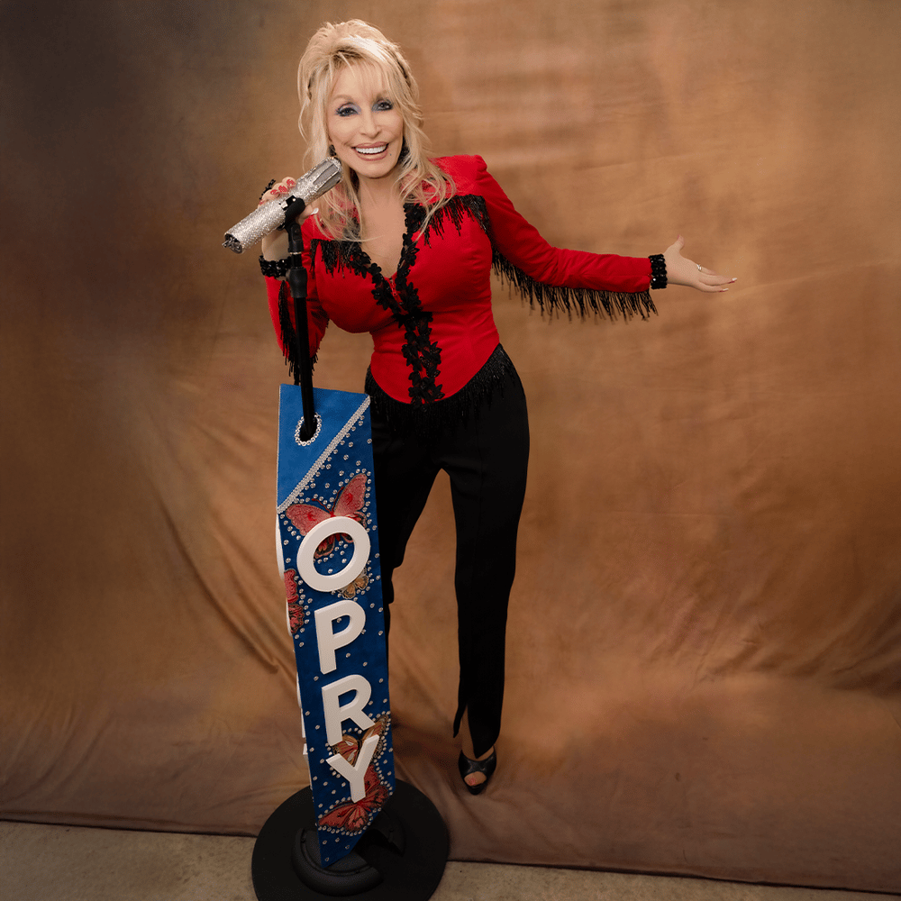 Dolly Parton in a red fringed top and black pants, smiling and holding a microphone, next to a blue "OPRY" stand with butterflies.