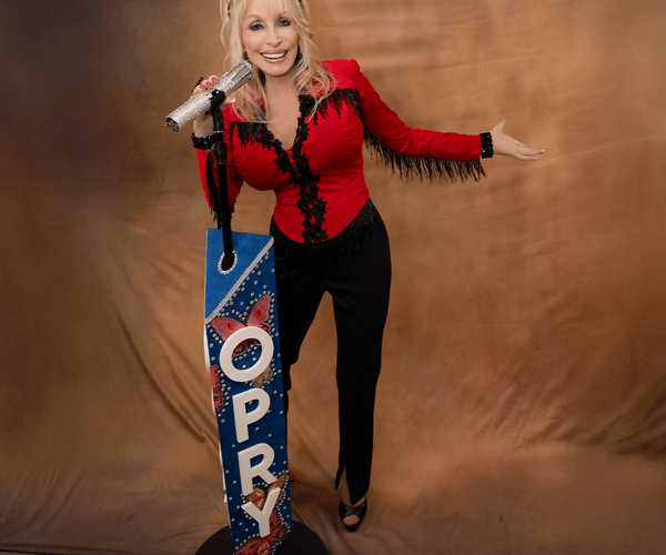 Dolly Parton in a red fringed top and black pants, smiling and holding a microphone, next to a blue "OPRY" stand with butterflies.