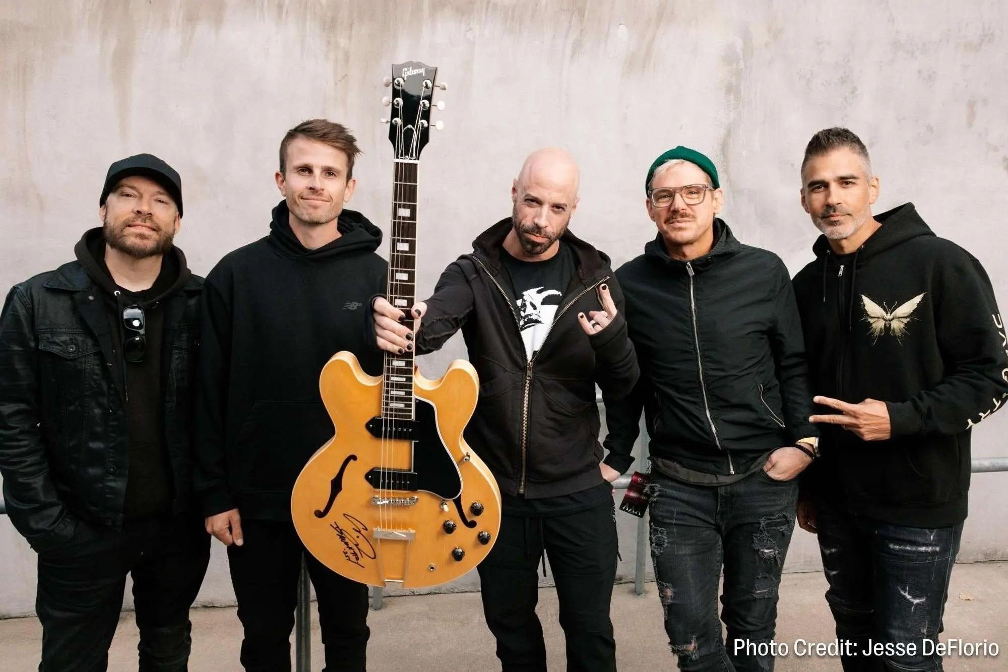 Five men posing together, one holding a tan electric guitar.