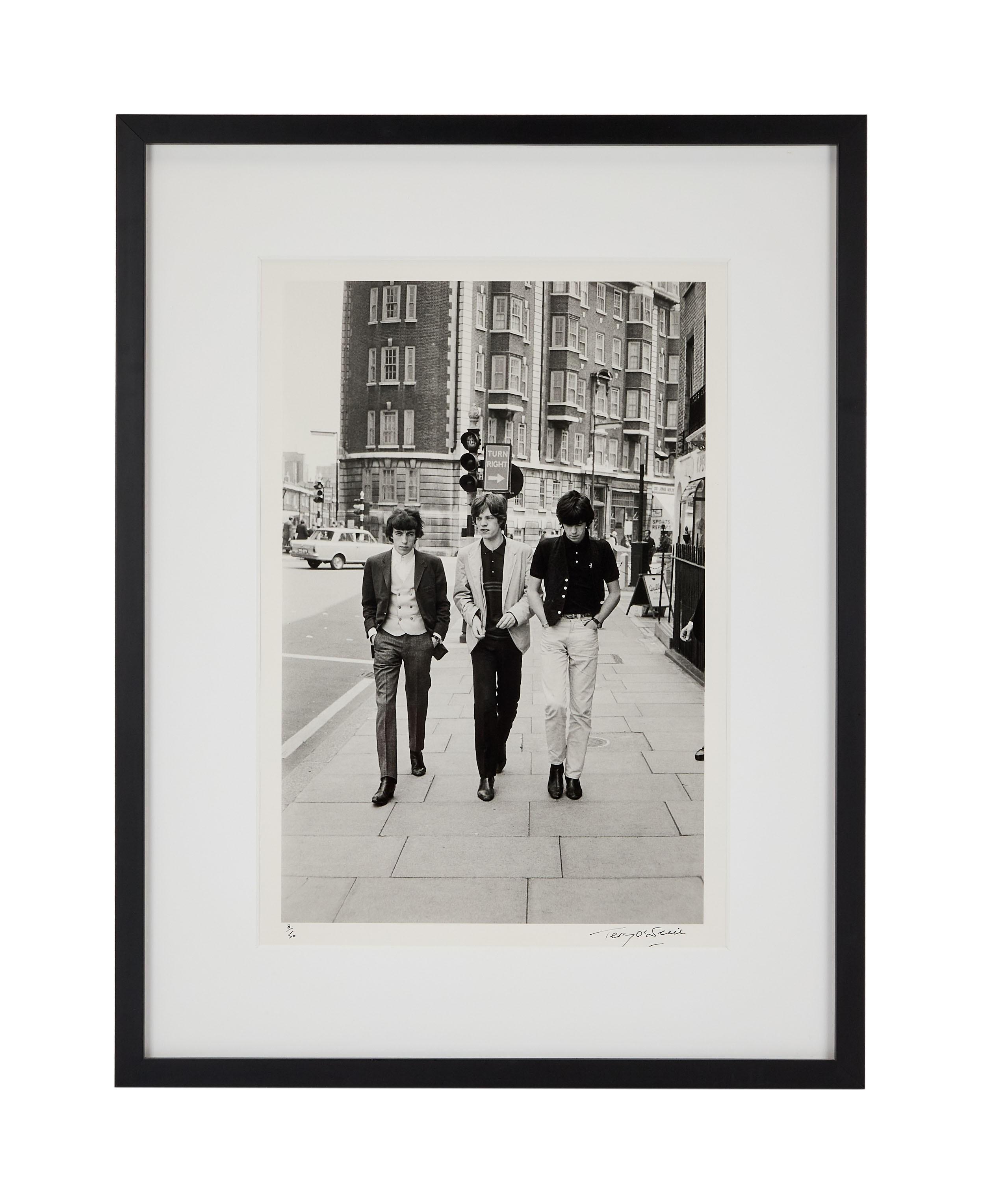 a black and white photo of the rolling stones walking down a sidewalk .