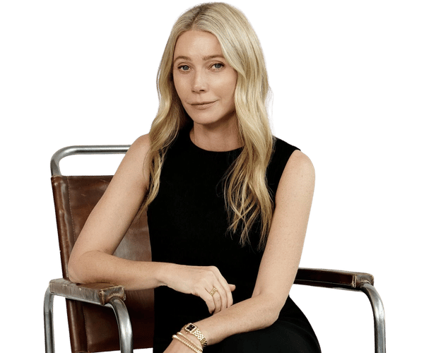A woman with long blonde hair in a black dress sits in a brown leather chair, wearing gold jewelry.