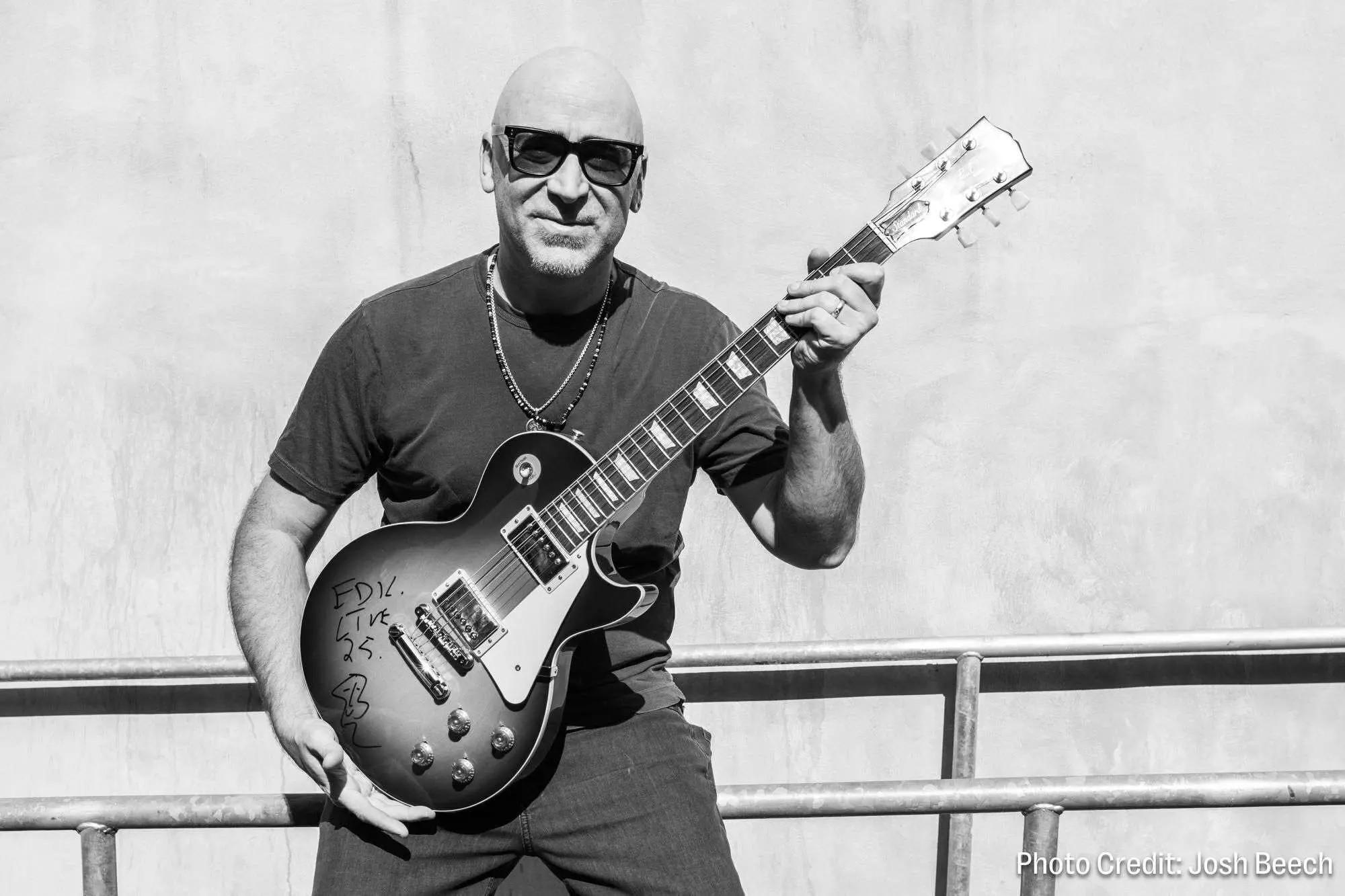 Black and white image of a bald man in sunglasses holding an electric guitar with writing on it.