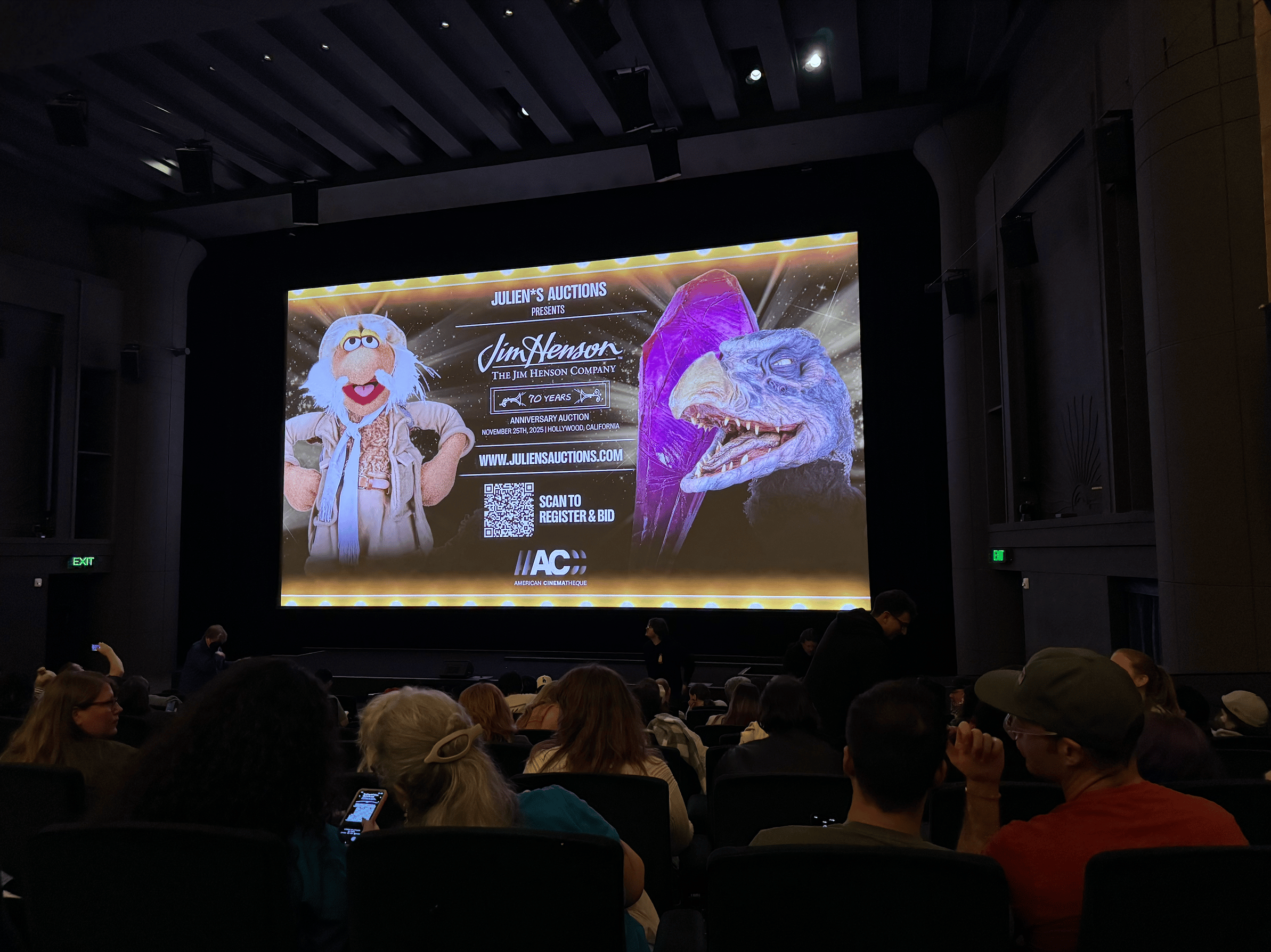 An audience in a dark theater watches a screen displaying a Julien's Auctions ad for Jim Henson, featuring Dr. Bunsen Honeydew and a Skeksis.