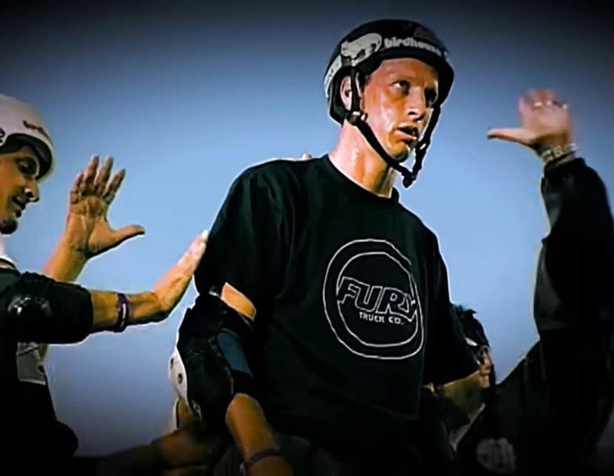 Tony Hawk, sweaty in a helmet and black t-shirt, is patted on the shoulders by two people.