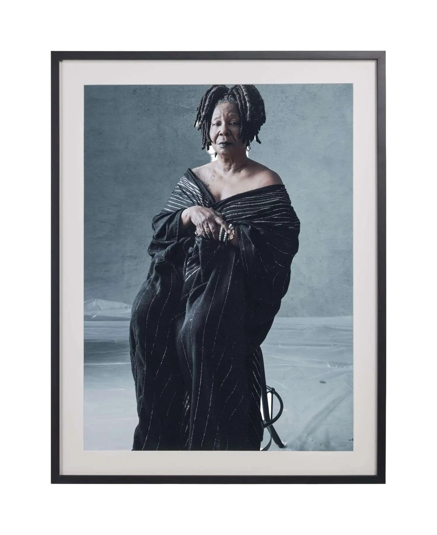 Framed portrait of Whoopi Goldberg seated, wearing a dark off-the-shoulder striped gown, against a grey background.