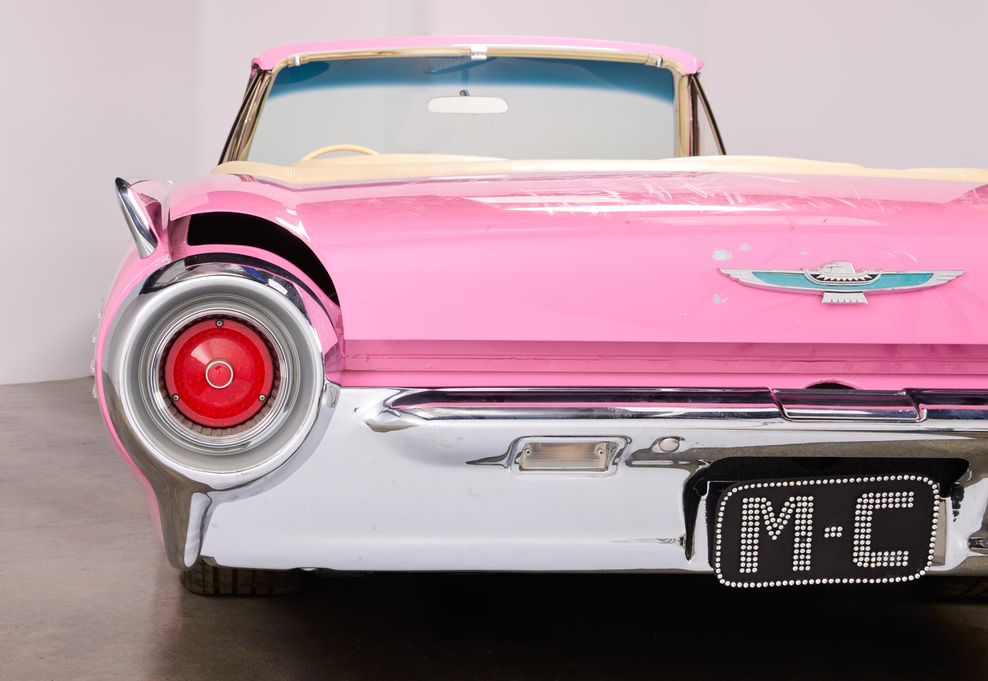 Rear view of a pink convertible car with a large round tail light, chrome bumper, and a sparkling "M-C" license plate.