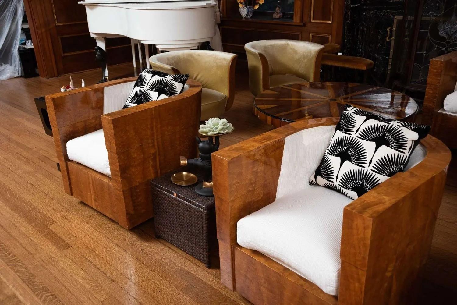 Art deco living room with two wooden armchairs, white cushions, patterned pillows, a round coffee table, and a white grand piano.