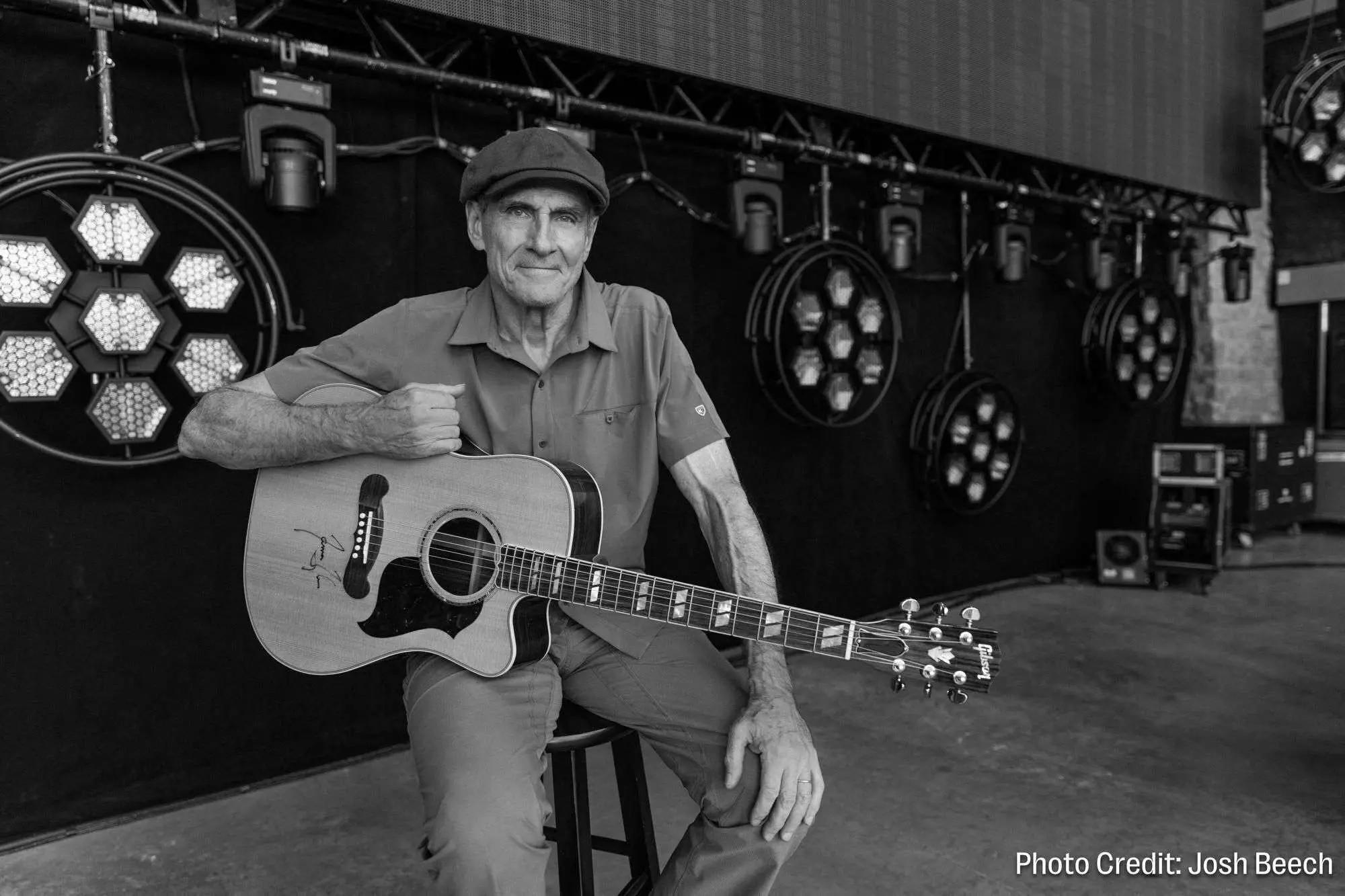 Black and white photo of an older man in a cap, smiling while holding an autographed acoustic guitar on a stage with lights.
