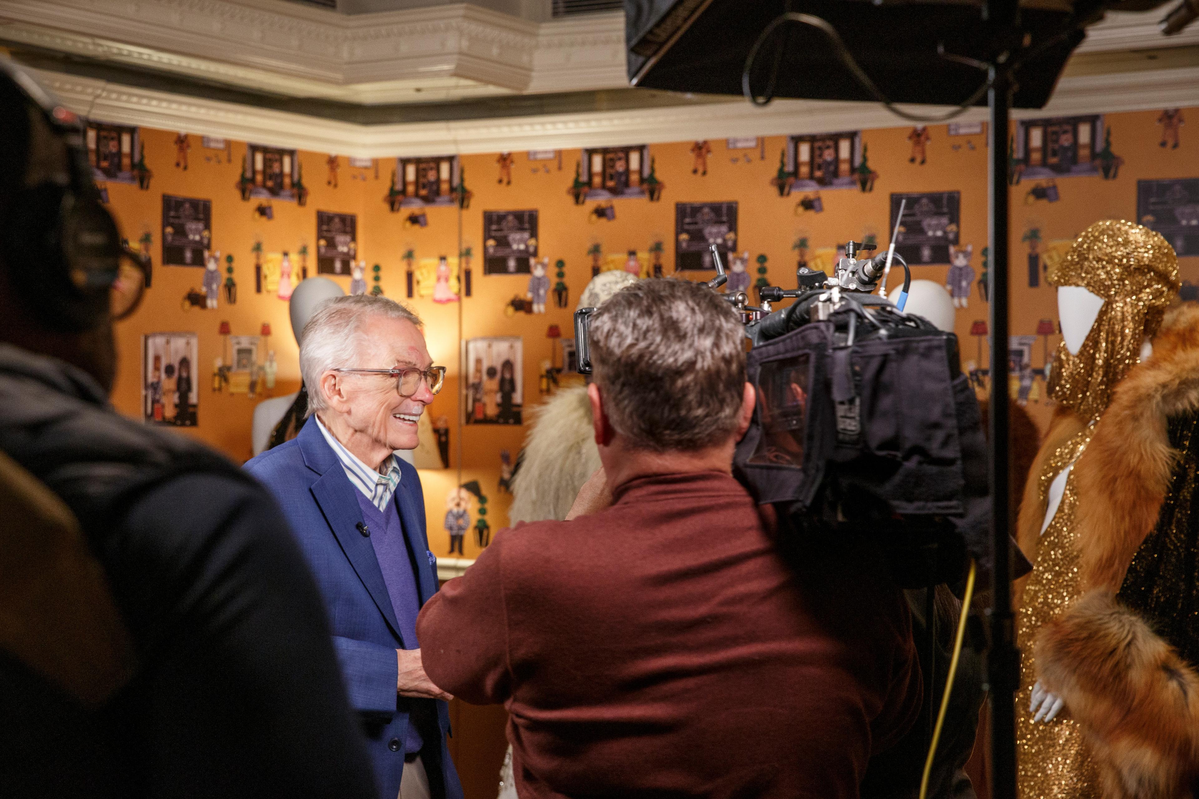 a man in a suit is talking to a cameraman in front of a wall with pictures on it .